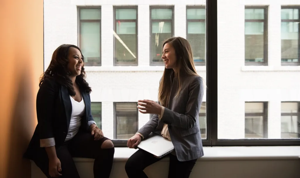 Mentoring vs Coaching: 2 Big Differences and Use Cases 6 An image of two women sitting in a window sill in an office. They are discussing important topics, such as mentoring vs coaching, while also laughing together.